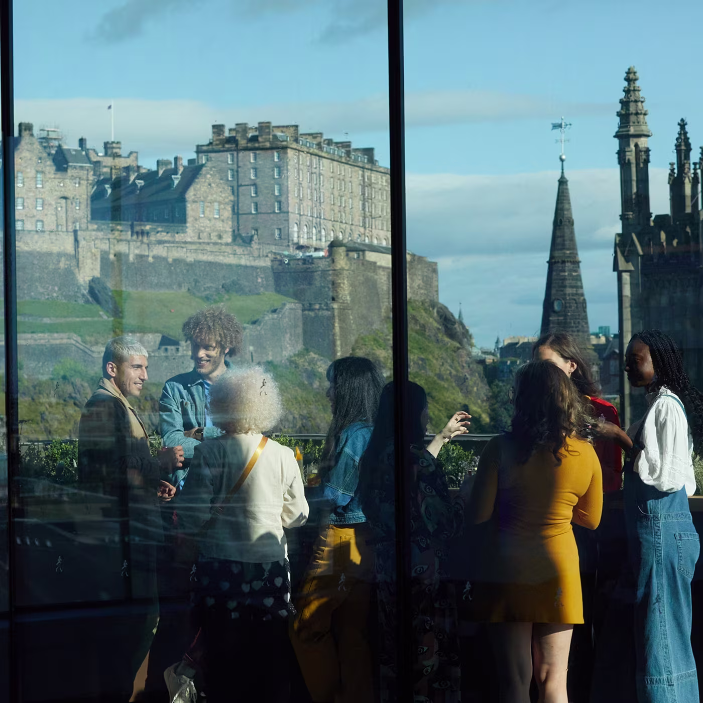 A group of people talk outside, reflected in glass, with an old castle and towers in the background.
