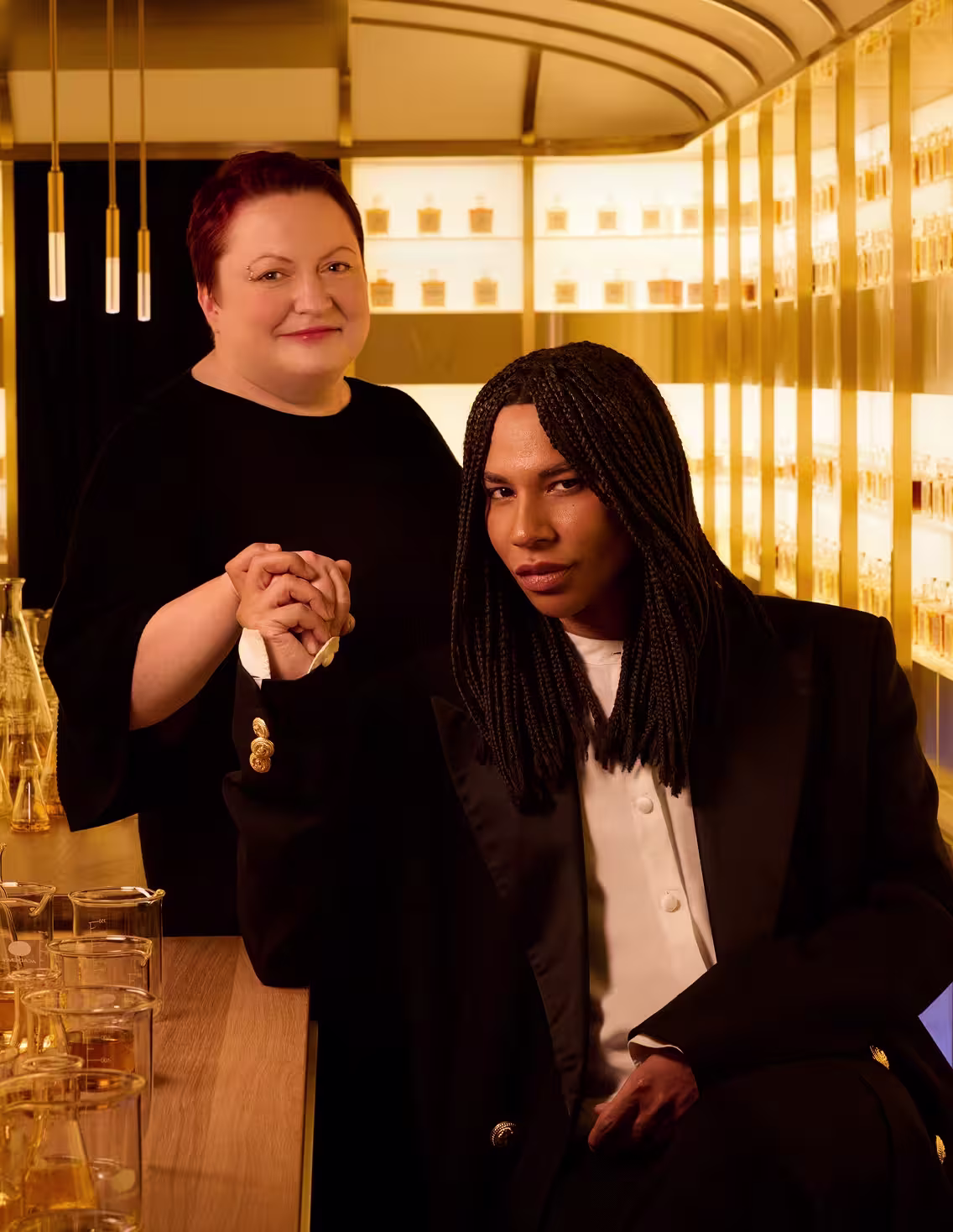 Two people pose in an elegant, golden-lit room lined with perfume bottles. One stands smiling in black, while the other, with braided hair and wearing a black suit, sits and holds the others hand, looking confidently at the camera.