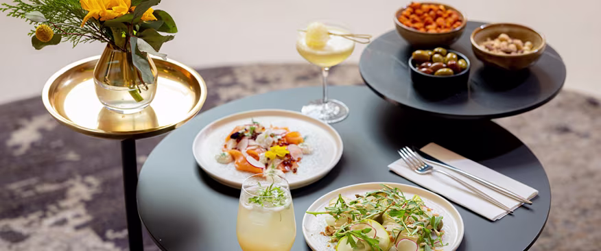 Une table élégante avec deux assiettes gourmandes, un cocktail, une limonade, des coupes d'olives et de noix, et un vase de fleurs jaunes sur un plateau doré. Les couvertures et les serviettes sont soigneusement disposées à côté des assiettes.