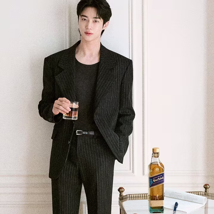 A person in a pinstripe suit holds a drink beside a table with a whiskey bottle and a notepad.
