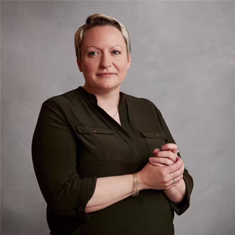 La Dra. Emma Walker, con cabello rubio corto y una camisa abotonada de color verde oscuro, se encuentra sobre un fondo gris liso y mira a la cámara con las manos juntas.