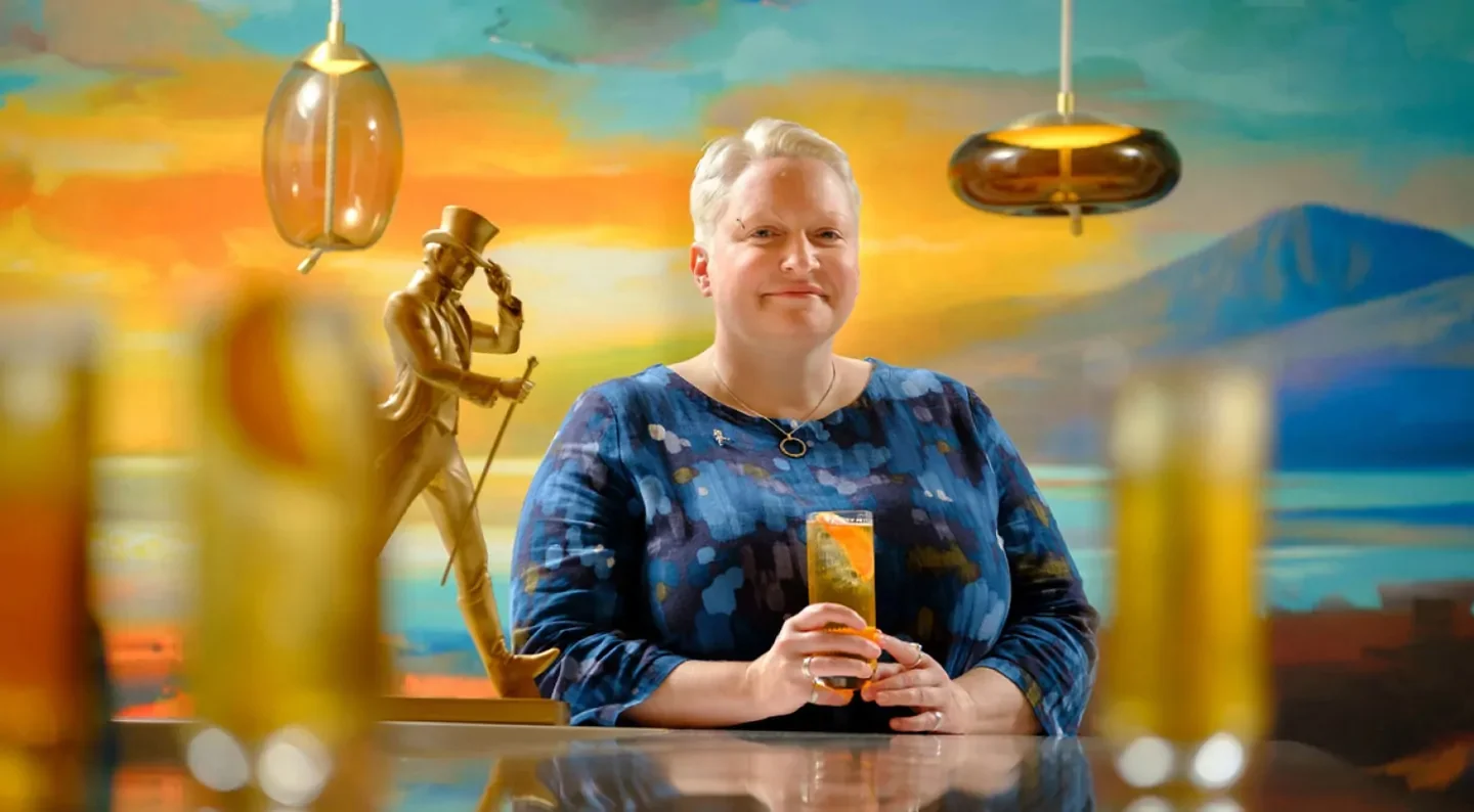 A person with short blonde hair, wearing a blue patterned dress, is holding a cocktail and standing at a bar. Behind them is a colorful mural and gold statue under modern hanging lights.