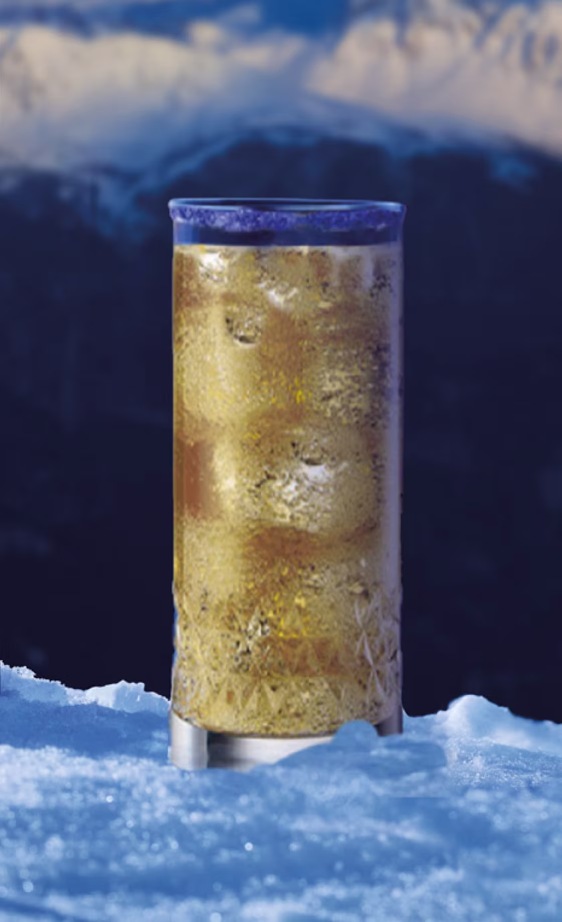 ぼやけた山々を背景に、金色の炭酸飲料が入った背の高いグラスが雪の上に置かれている。グラスは霜が降りたような外観で、冷たい飲み物であることを示唆している。.