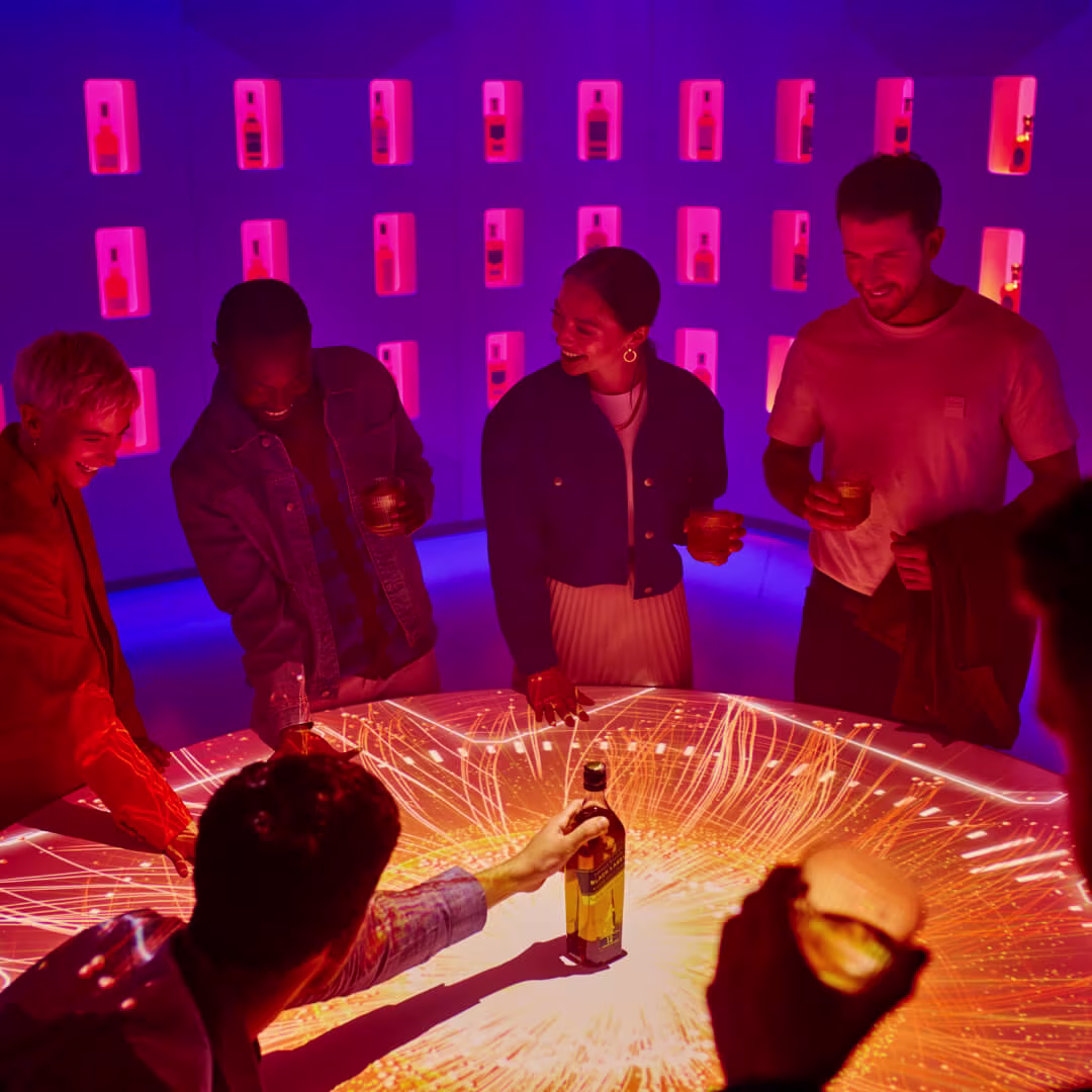 A group of people in a dim, colorful room watch as a man touches smoke coming from a small bowl held by a woman. The atmosphere is lively and mysterious, with a ceiling decorated with flowers and greenery.