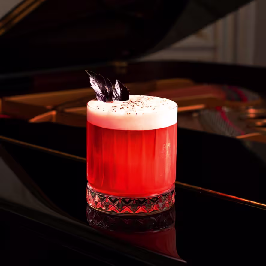 A vibrant red cocktail with a frothy top, garnished with dark leaves, sits in a textured glass on a glossy black piano. Soft light highlights the drink’s rich color and elegant presentation.