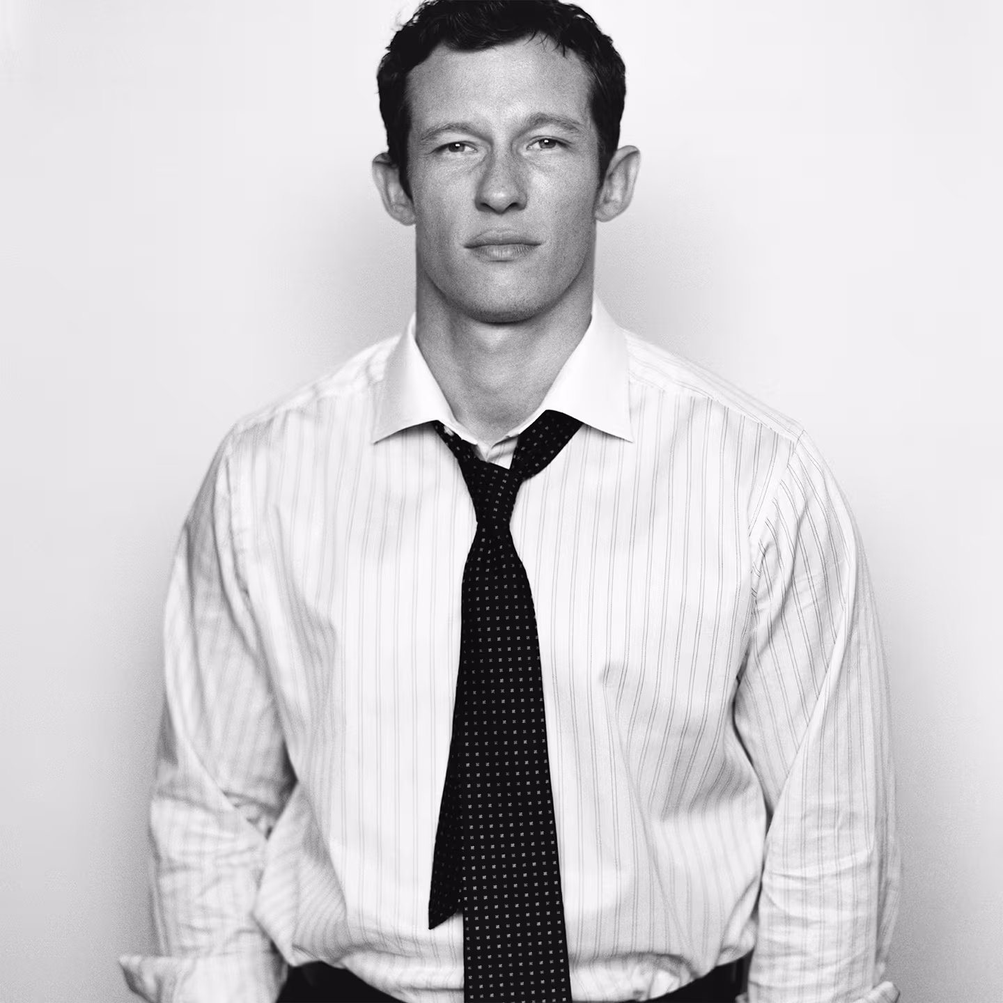 Callum Turner in a striped dress shirt and loose tie standing against a plain background, looking at the camera.