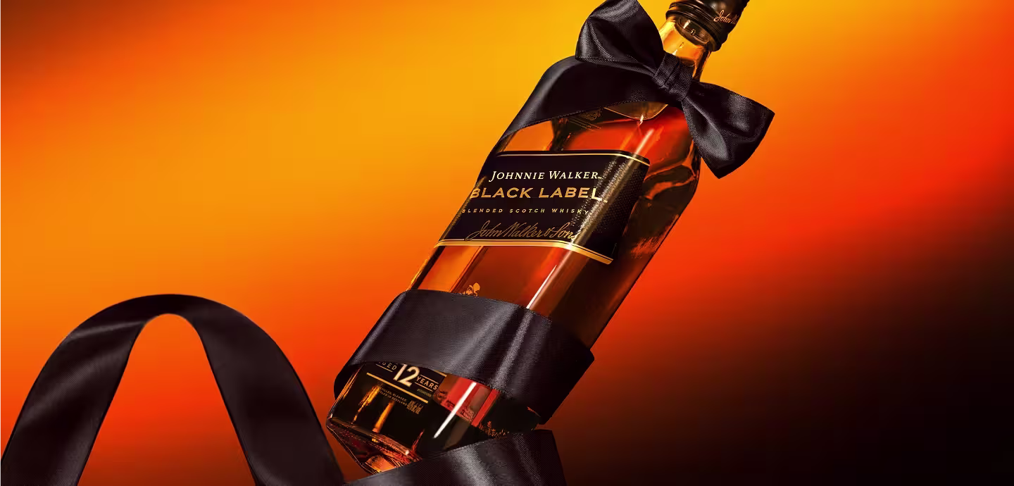 Eine Flasche Johnnie Walker Black Label Whisky ist mit einem schwarzen Satinband umwickelt, das zu einer Schleife gebunden ist, und steht vor einem leuchtend orange-schwarzen Hintergrund mit Farbverlauf.
