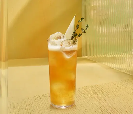De Zoete Fruit Lentebeker. Een hoog glas gevuld met een amberkleurige ijsdrank, afgedekt met een schuimlaagje, een takje tijm en een dun wit schijfje als garnering. Het glas wordt op een lichtgekleurd oppervlak met een structuurpatroon geplaatst.