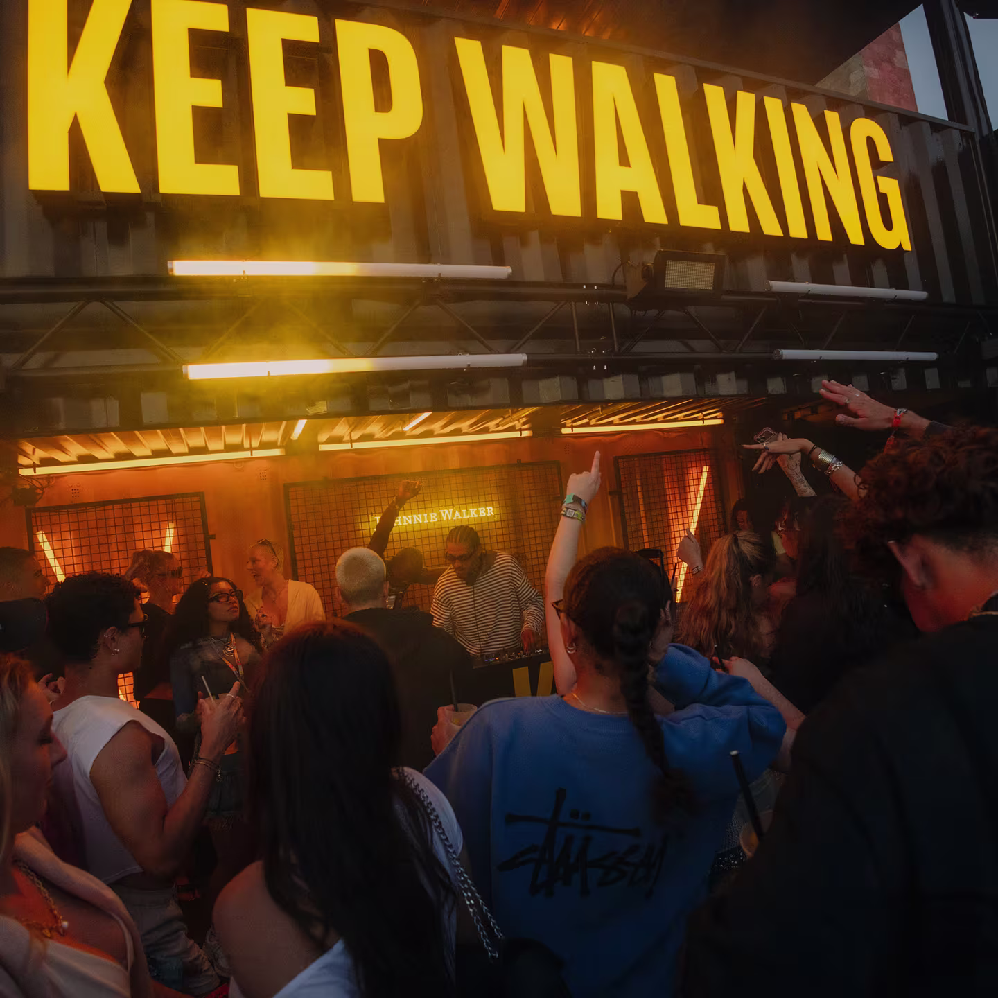 Eine tanzende Menge unter einem großen gelben Keep-Walking-Schriftzug an einer Festivalbar.