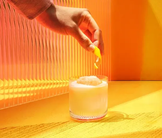 Mão colocando uma casca de limão em um copo de bebida gelada sobre uma superfície amarela brilhante. A luz do sol projeta sombras através do vidro canelado, criando uma atmosfera quente e vibrante.
