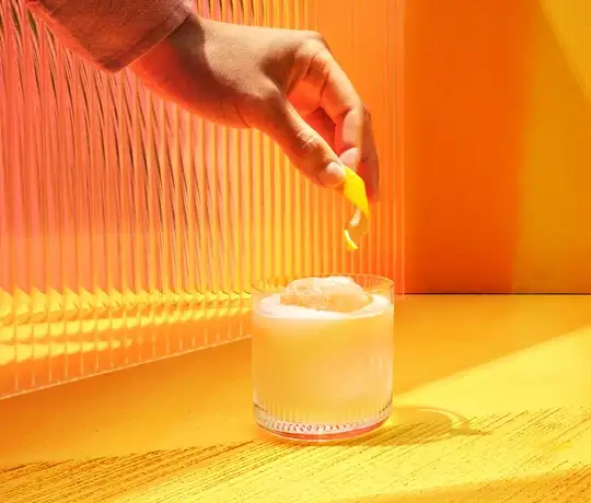 The Smooth & Floral Old Fashioned. A hand holds a lemon peel over a glass filled with a creamy white beverage and ice. The background features bright yellow and orange hues with a textured wall, creating a warm and sunny ambiance.