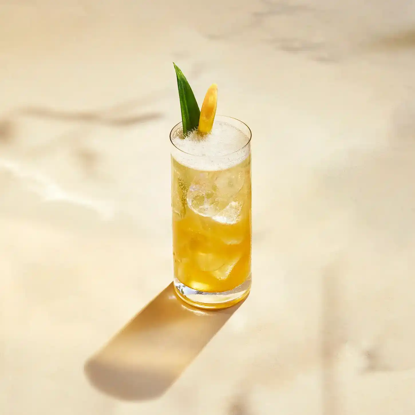 A tall glass of yellow, iced cocktail garnished with a lemon wedge and a sprig of green foliage. 
