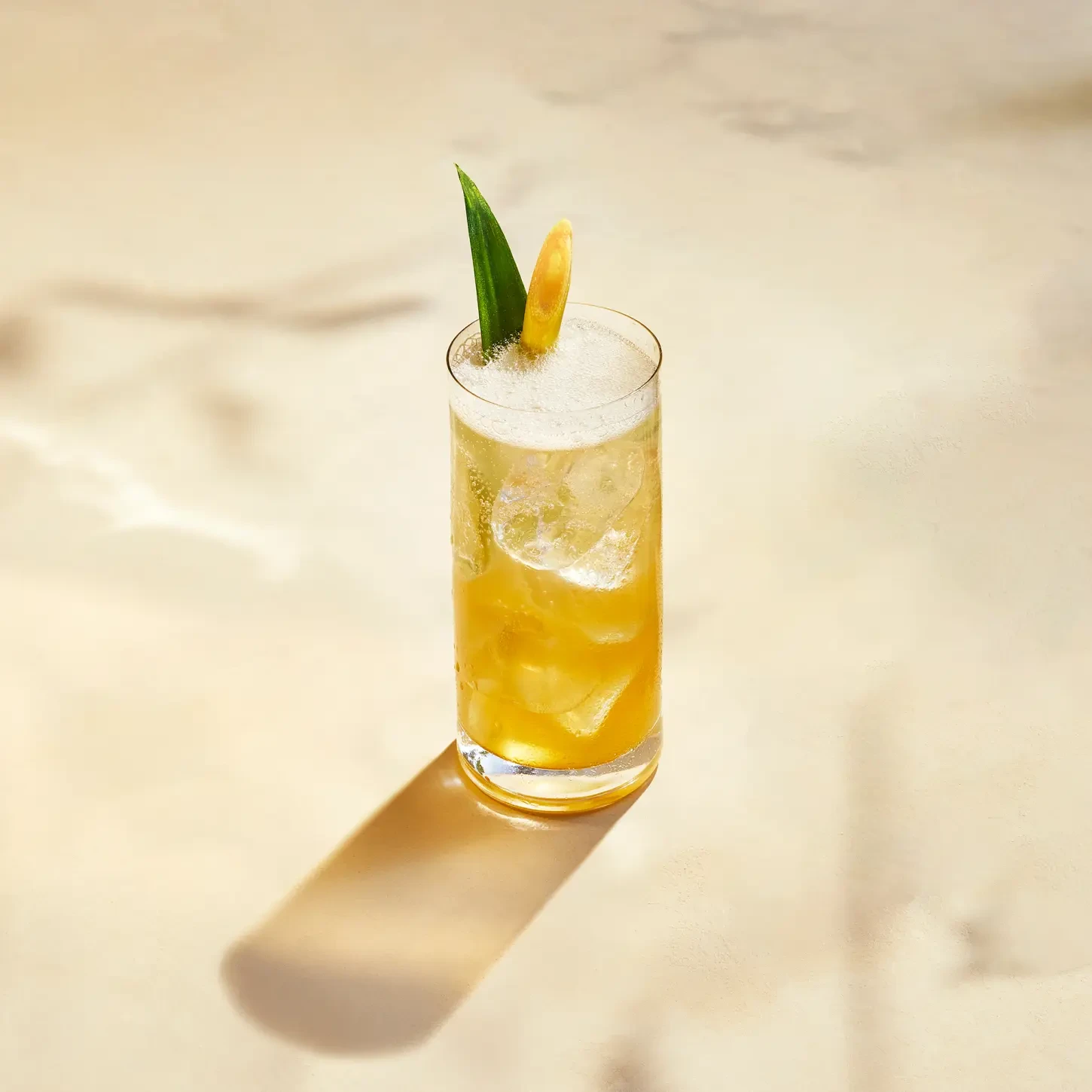 A tall glass filled with a yellow tropical drink, garnished with a pineapple leaf and citrus slice. The drink is on a light surface, creating a soft shadow. Ice cubes are visible in the drink, and foam is on the top.