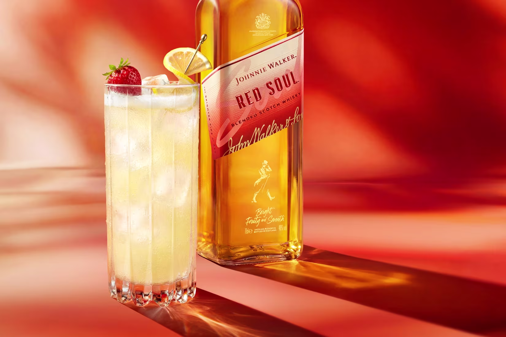 Sklenice koktejlu s ledem, plátkem citronu a jahodou vedle láhve whisky Johnnie Walker Red Soul.