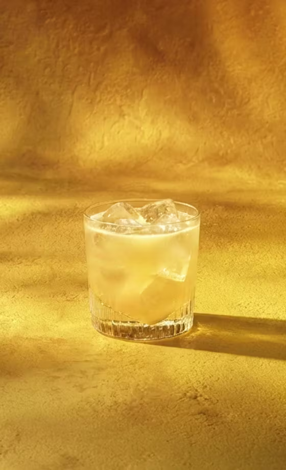 A glass filled with a light-colored drink and ice cubes sits on a textured golden-yellow surface, with warm light casting soft shadows.