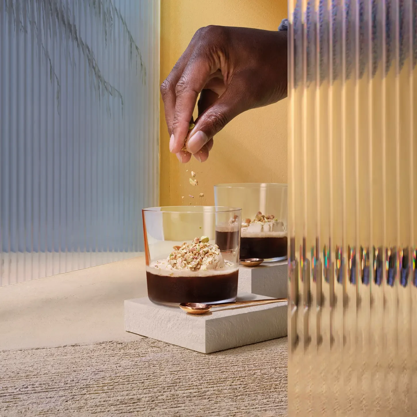 砕いたナッツをホイップクリームをトッピングしたコーヒーグラスに振りかけている手。背景はテクスチャーのあるベージュ色の表面と右側の金色の半透明のパネルで構成され、柔らかな自然光がシーンを引き立てています。.
