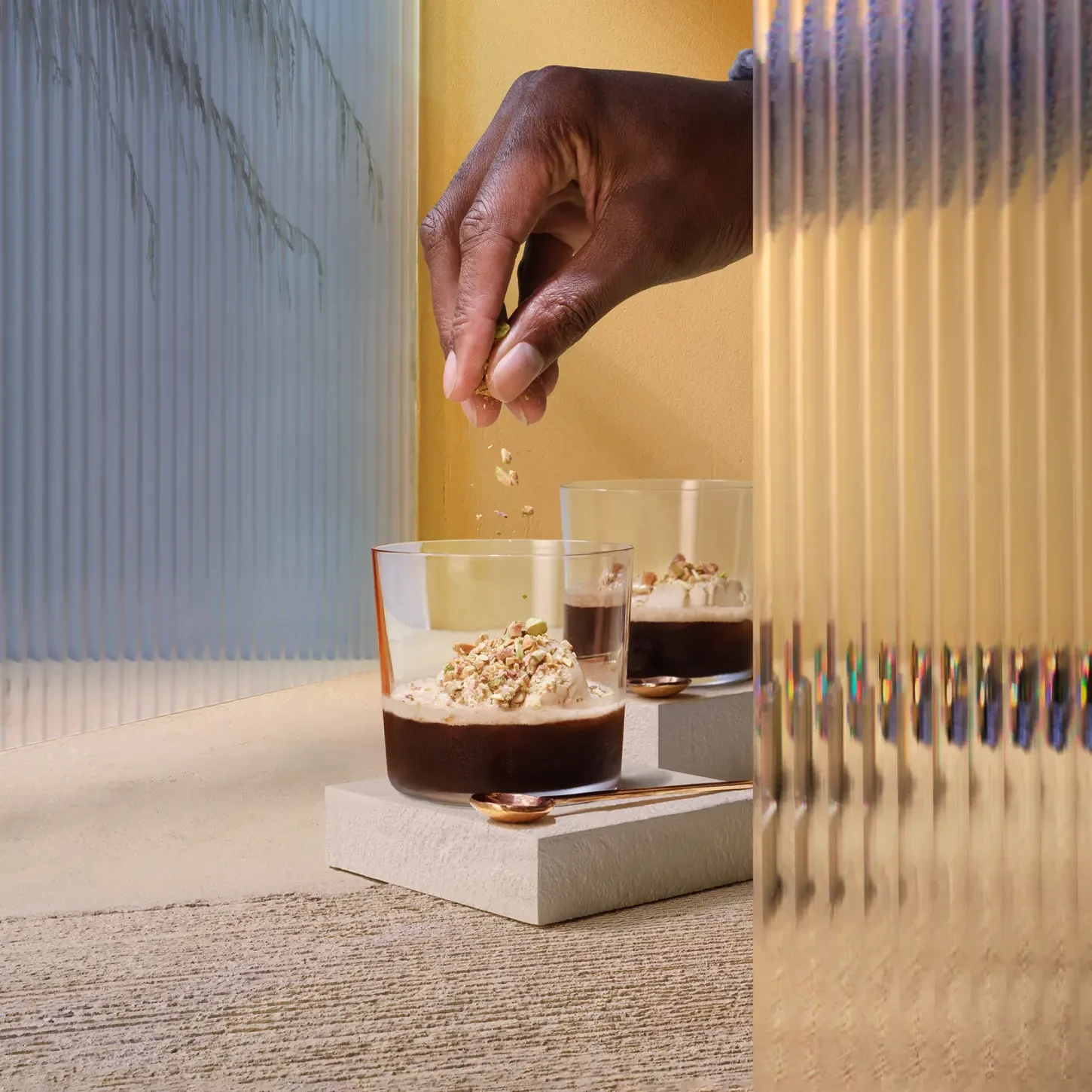 The Sweet & Nutty Affogato. A hand sprinkles toppings onto a dessert in a glass. The dessert appears creamy and is placed on a white surface. Another similar dessert glass is in the background. The setting features textured glass panels and warm lighting.