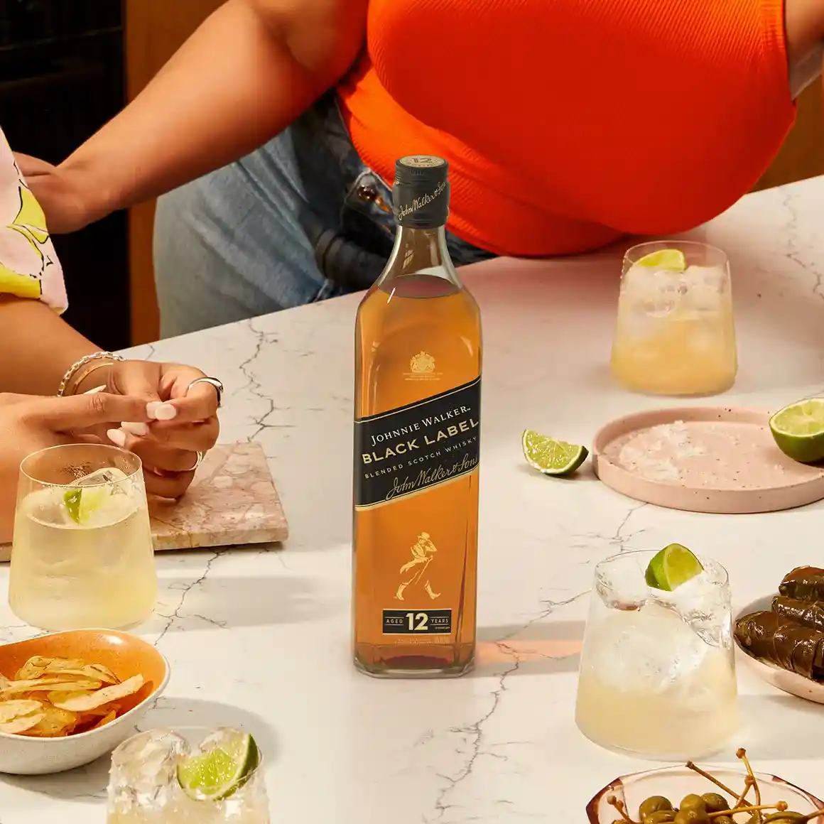 Sobre una encimera de mármol hay una botella de Johnnie Walker Black Label rodeada de cócteles con hielo y lima. Se ven parcialmente dos personas interactuando en la mesa. Cerca hay pequeños cuencos con aperitivos.