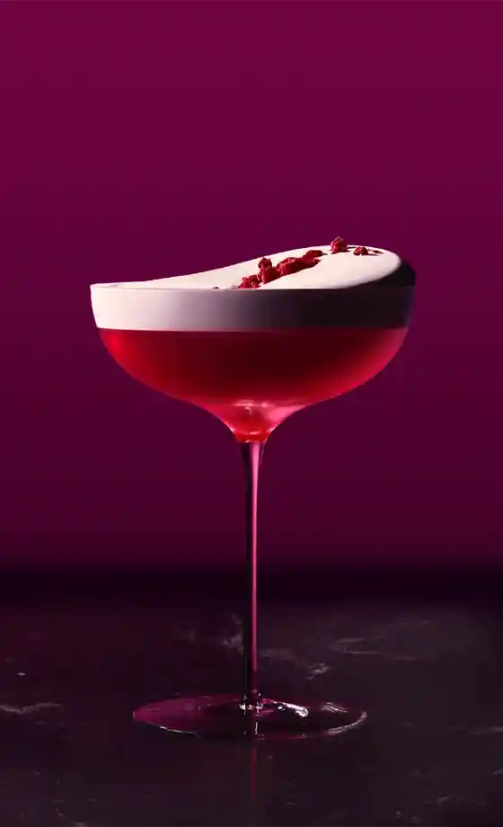 A vibrant pink cocktail in a coupe glass, topped with frothy white foam and decorated with red flakes, sits on a dark marble surface against a deep magenta background.