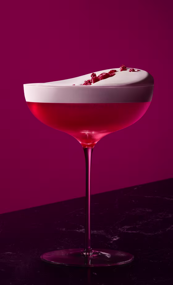 Un cóctel de color rosa vibrante en una copa coupé reposa sobre una superficie oscura, cubierto con una gruesa capa de espuma blanca y espolvoreado con guarnición roja, contra un fondo magenta intenso.