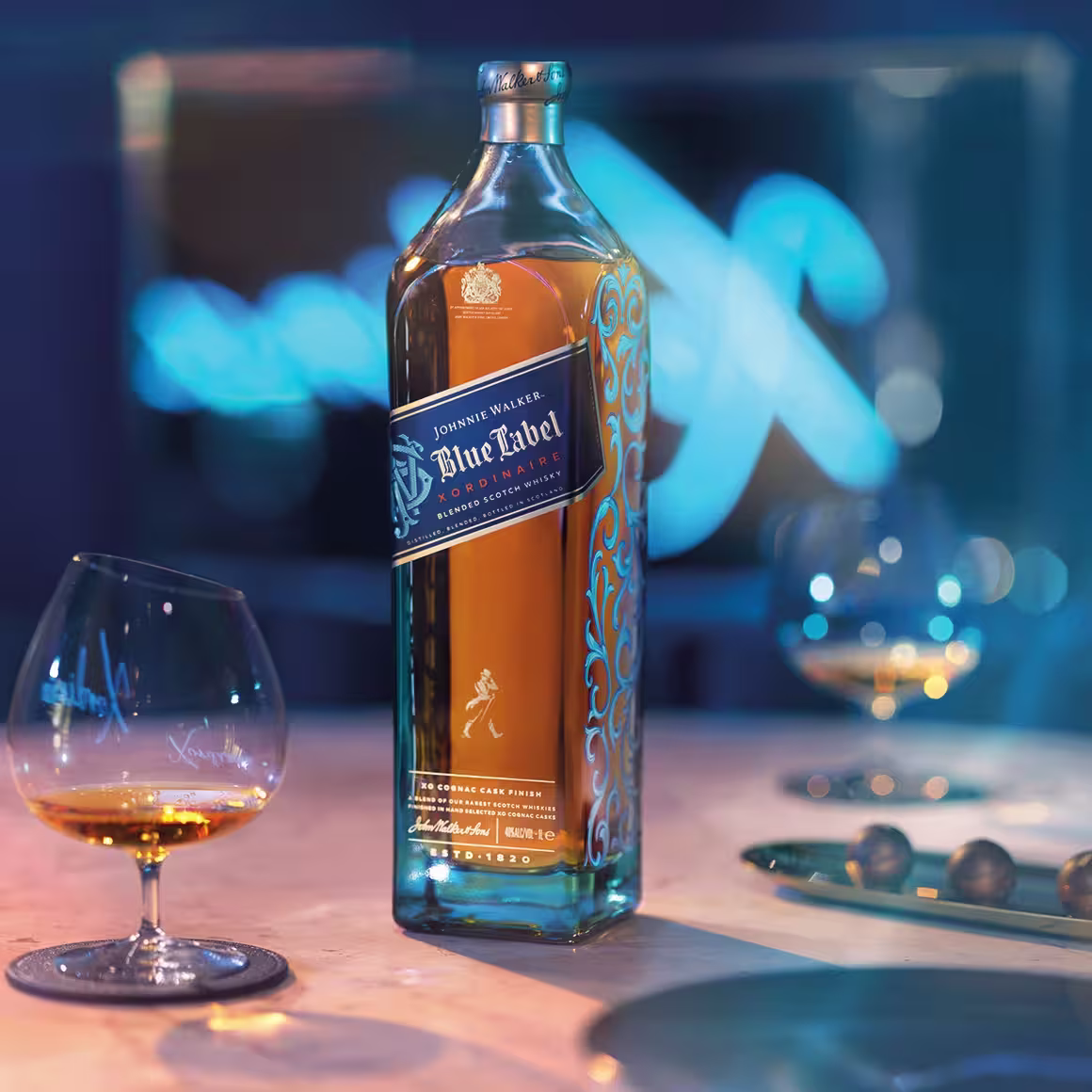 A bottle of Johnnie Walker Blue Label whisky stands on a table next to a glass of whisky, another glass in the background, and a blurred blue-lit backdrop. The scene has a sophisticated, elegant atmosphere.
