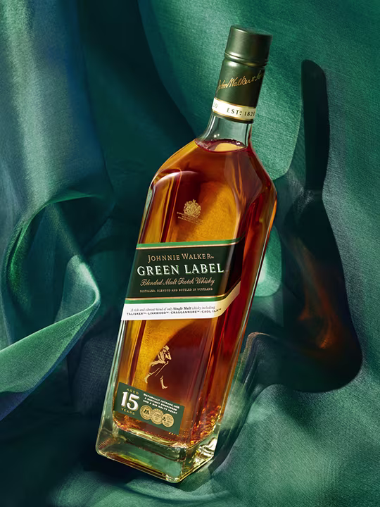 Бутилка Johnnie Walker Green Label смесено малцово шотландско уиски лежи върху фон с текстурирана тъкан. Етикетът показва, че е отлежал 15 години. Бутилката хвърля сянка, а материята има нюанси на зелено и синьо.
