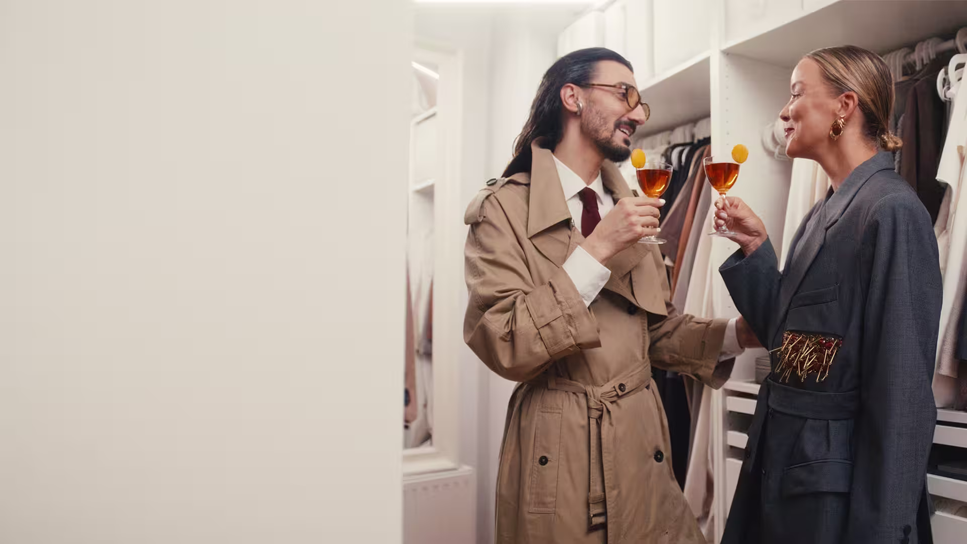 Zwei Menschen in stylischen Trenchcoats stehen lächelnd in einem begehbaren Kleiderschrank und stoßen mit Orangencocktails an, umgeben von ordentlich geordneter Kleidung und Accessoires.