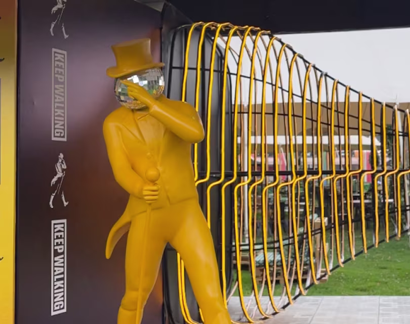 Una estatua amarilla con sombrero de copa y bastón se encuentra frente a una gran escultura que parece una botella tumbada. La estructura se encuentra dentro de un arco decorativo con la frase «KEEP WALKING».