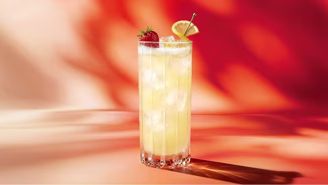 Hohes Glas mit eisgekühltem, gelbem Getränk, garniert mit einer Zitronenscheibe und einer Erdbeere, auf orange-rotem Hintergrund.