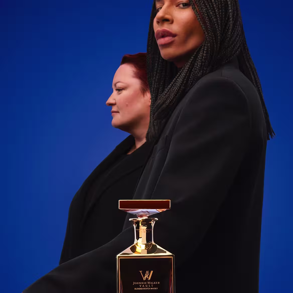 Two people in black jackets smile and stand close together behind a shiny black table. A gold perfume bottle sits on the table, reflecting their image. The background is plain white.