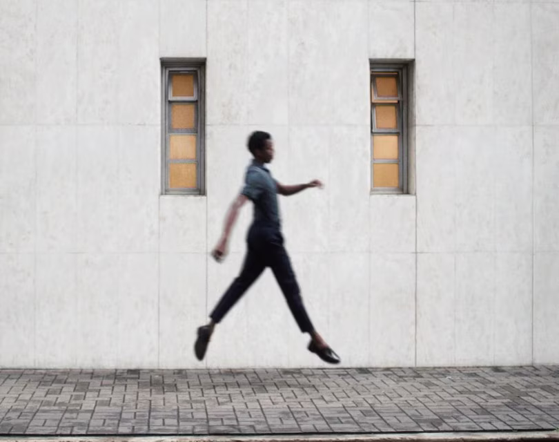 Un hombre es captado en el aire mientras salta sobre una acera frente a un edificio con ventanas estrechas. Otra persona camina delante en dirección opuesta, creando una sensación de movimiento y contraste contra la estática pared.