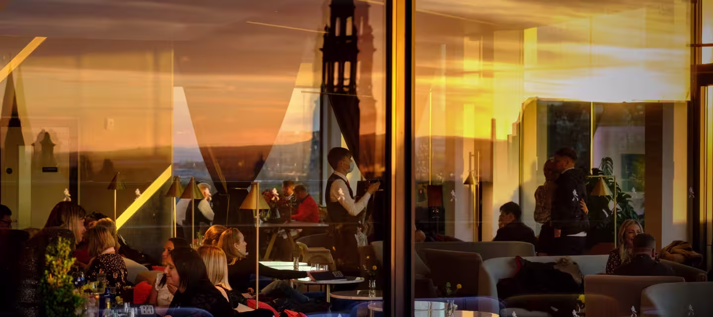 People sit and socialize in a stylish indoor lounge as warm sunset light streams through large windows, reflecting cityscape and architecture outside. A waiter stands in the center, and some guests are silhouetted against the golden light.