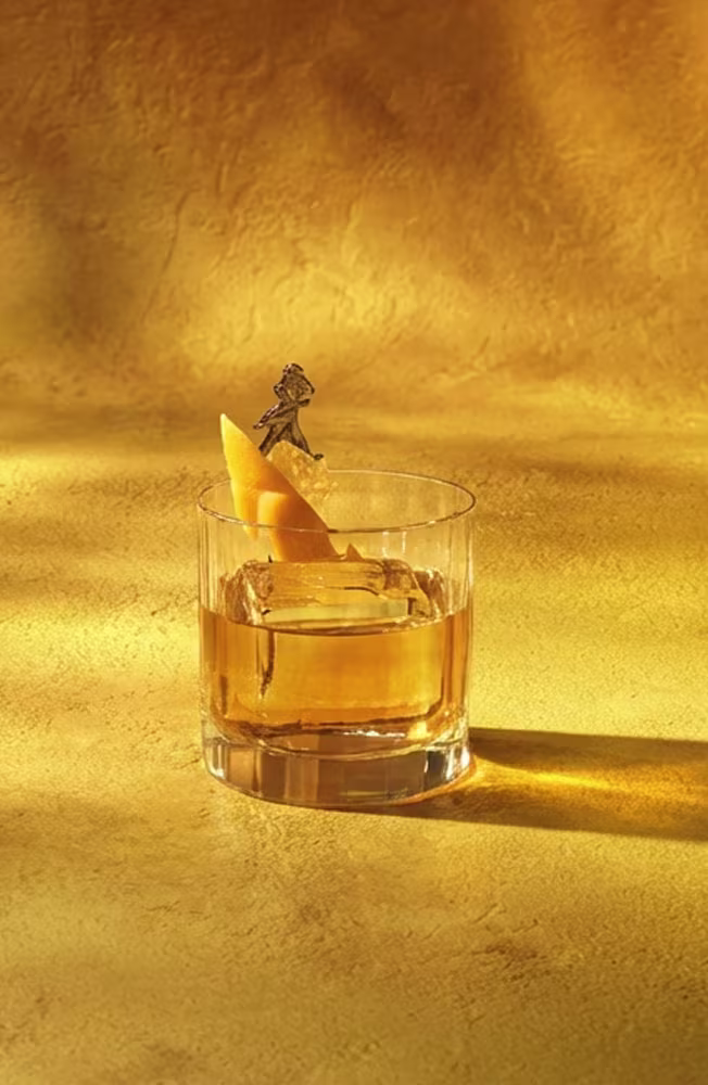 A glass of whiskey with a large ice cube and an orange peel garnish, held by a decorative stick, sits on a sunlit yellow surface against a matching textured background.