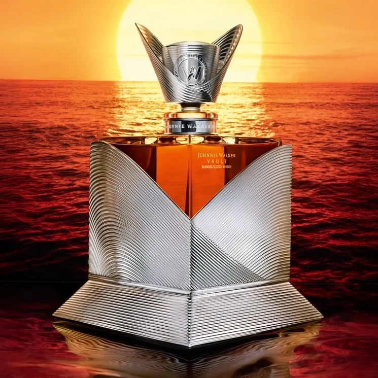 A luxurious Johnnie Walker whiskey bottle with an intricate silver design is set against a vibrant sunset over the ocean, with golden sunlight reflecting off the waves and the bottle’s metallic surface.