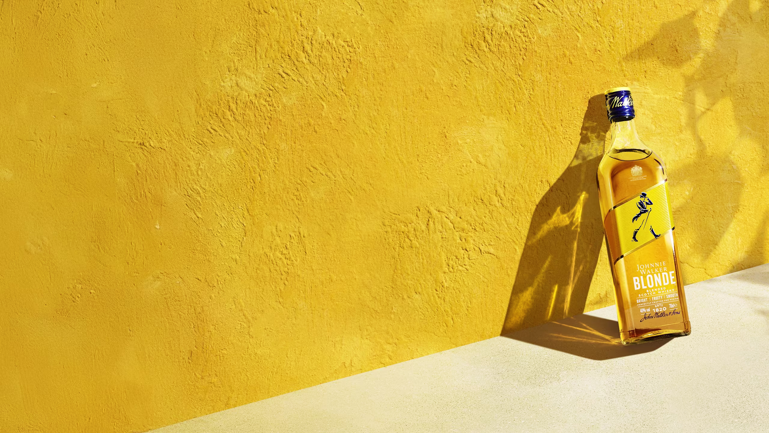 A bottle of blonde whiskey stands upright against a vibrant yellow background, casting a shadow. The label features a walking figure in blue. Sunlight highlights the textured surface.