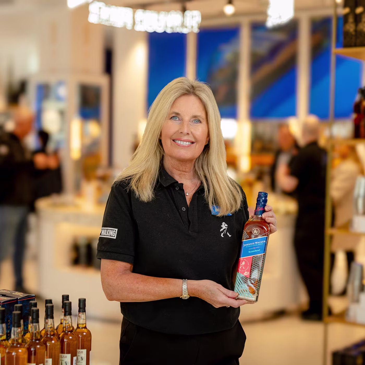 Smiling woman in a black polo shirt holds a bottle of whisky in a well-lit store.