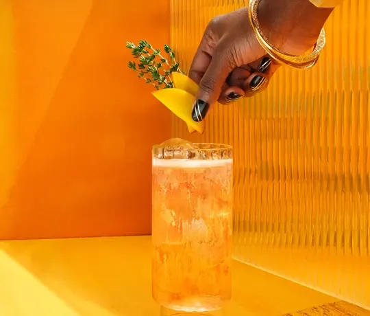 Une main avec des bracelets en or et du vernis à ongles noir garnit un grand verre de boisson à l'orange avec un quartier de citron et des herbes sur un fond jaune vif.