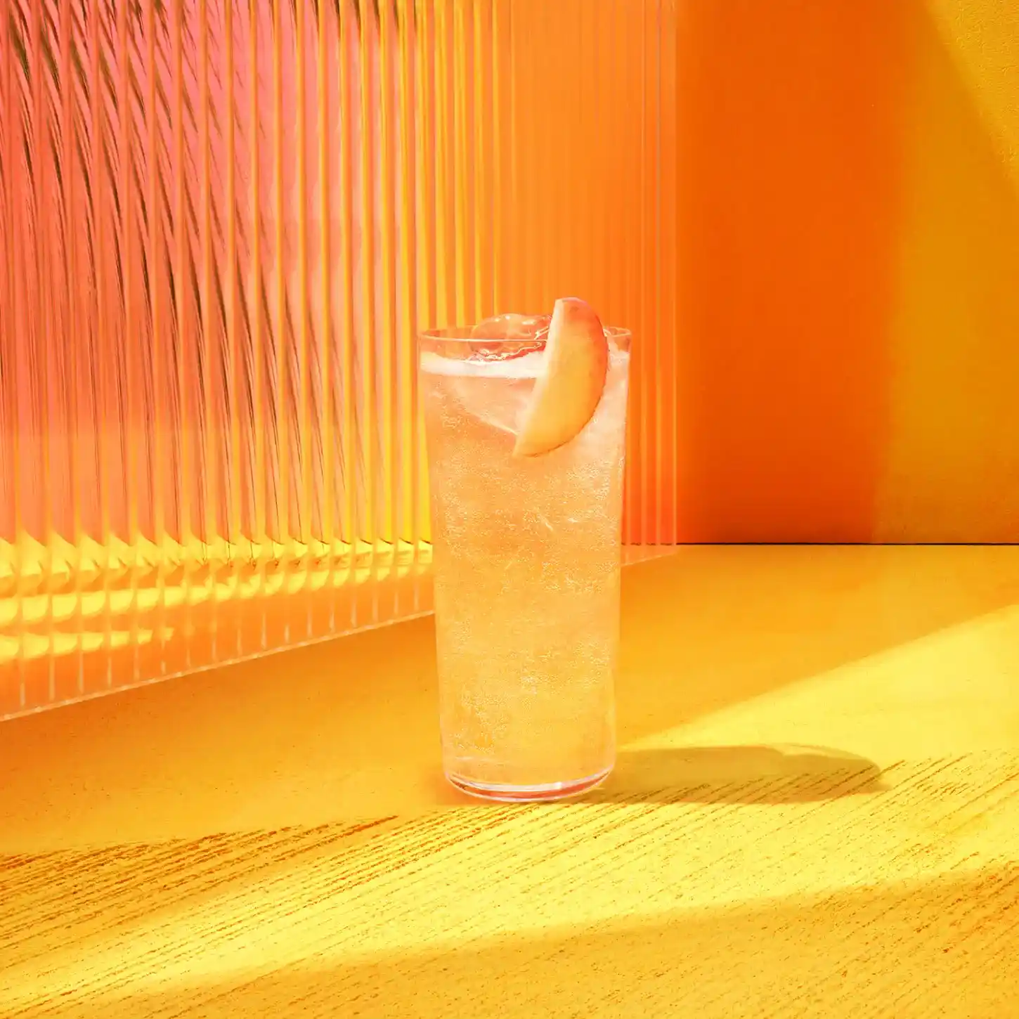 A tall glass of a light-colored drink with a peach slice garnish sits on a yellow surface in front of an orange and pink striped background. 
