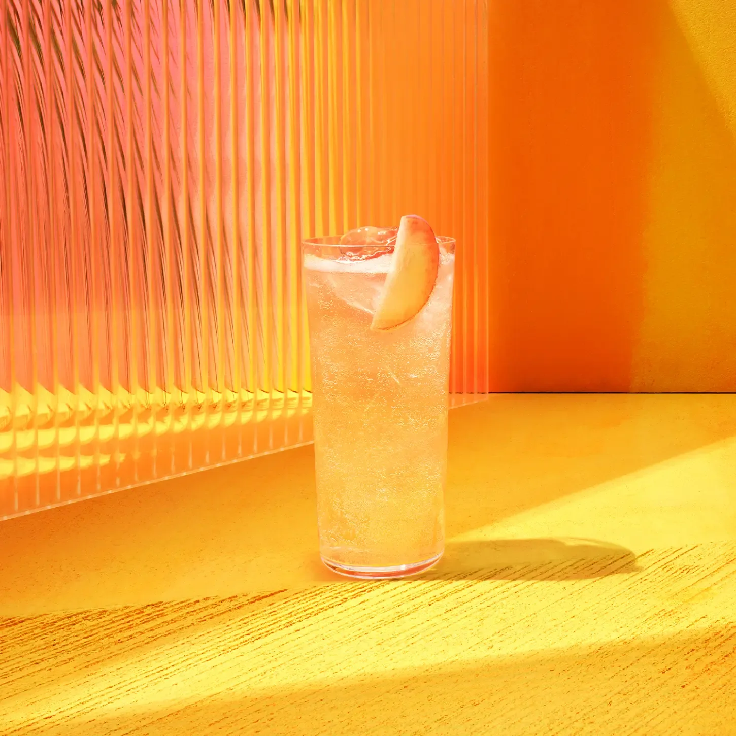 The Spiced Citrus Stone Fence. A tall glass with a peach-colored drink and ice, garnished with a peach slice. Its placed on a bright yellow surface with a textured, translucent pink backdrop. Sunlight casts warm reflections around the scene.