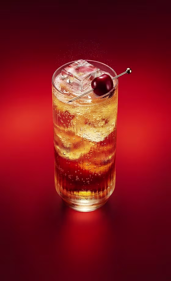  Un vaso alto de cóctel ámbar efervescente con hielo y una cereza como decoración, sobre un fondo rojo.