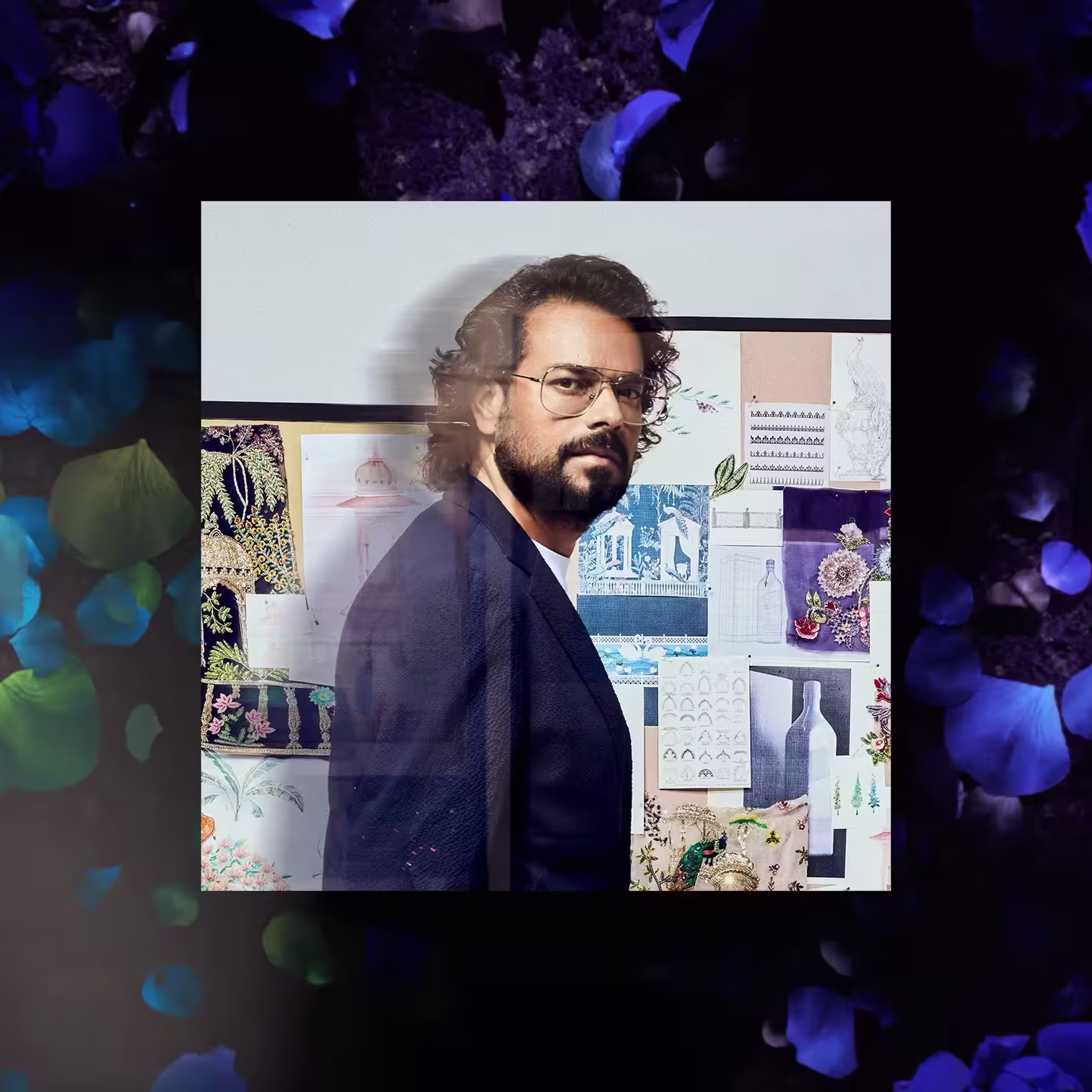 Portrait of fashion designer Rahul Mishra with long dark hair, glasses and a beard, wearing a navy jacket against dark floral imagery.