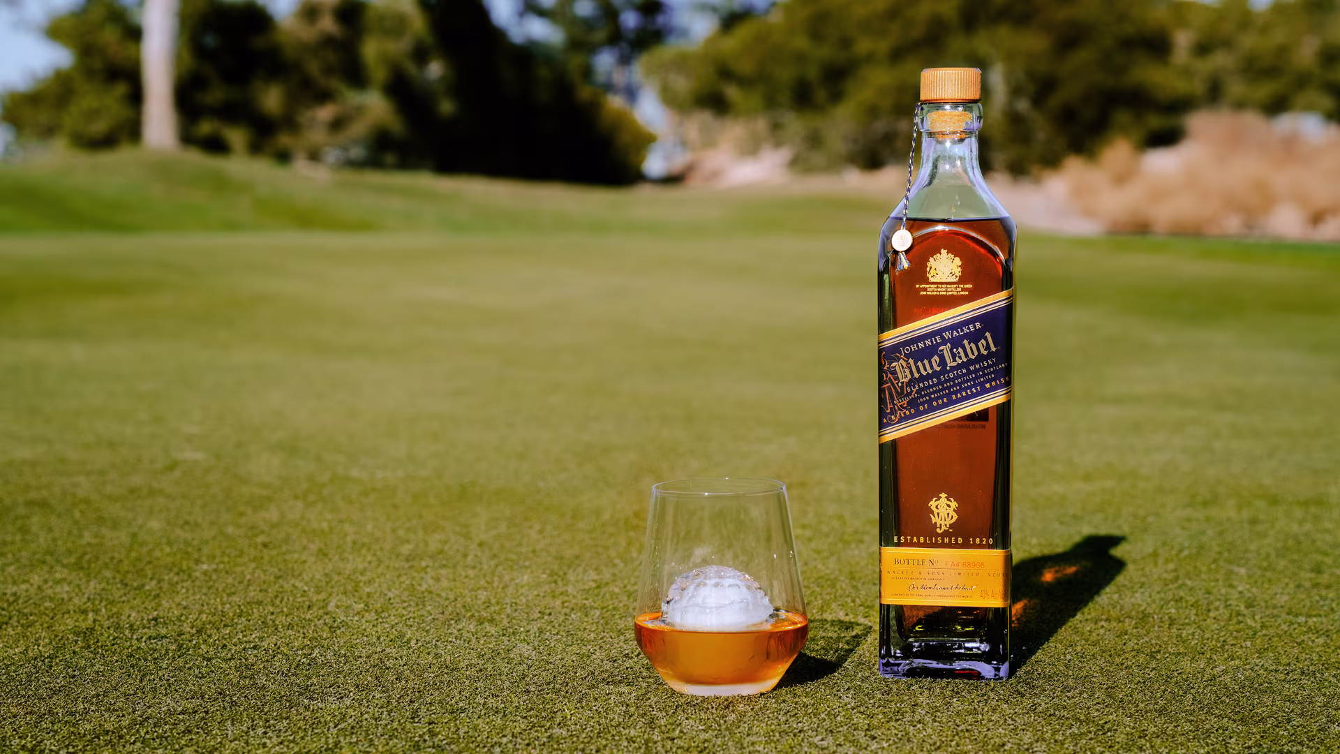 A bottle of Johnnie Walker Blue Label whisky and a glass with whisky and a golf ball sit on a green golf course with trees in the background.