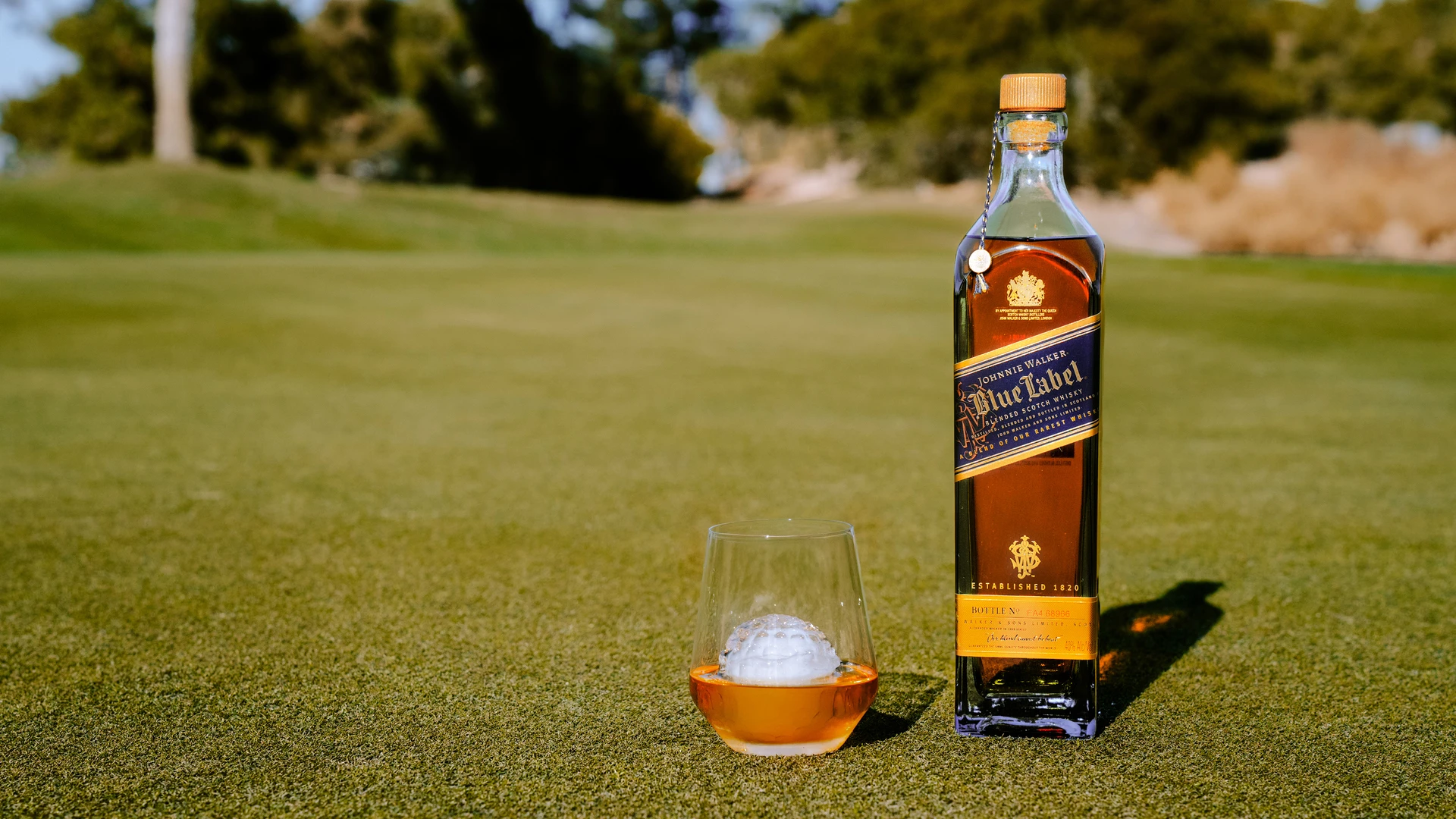 A bottle of Blue Label whiskey is on a golf course, next to a glass containing whiskey and a golf ball-shaped ice cube. The background features blurred trees and a clear sky.