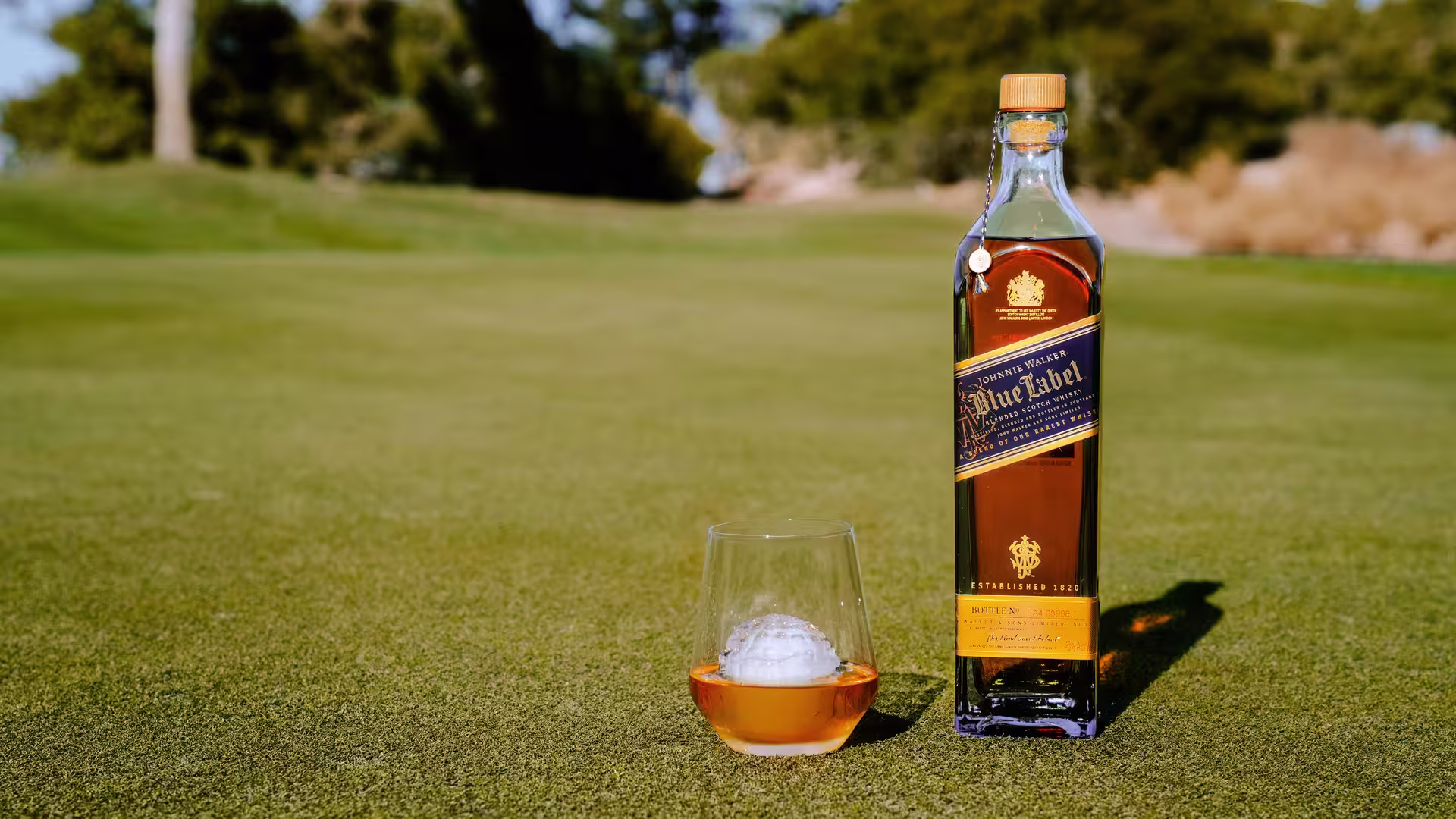 A bottle of whiskey labeled Blue Label stands on a grass surface next to a glass of whiskey containing a golf ball-shaped ice cube. The background is a blurred landscape with trees.