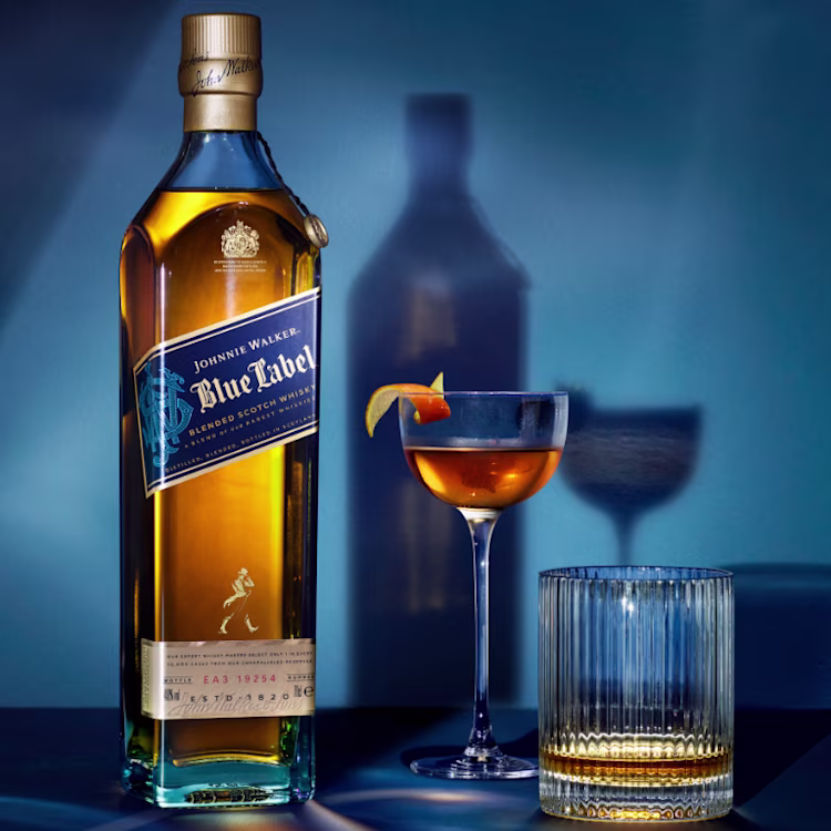 A bottle of Johnnie Walker Blue Label whiskey is displayed alongside a cocktail in a coupe glass with an orange twist garnish. A glass cup is seen nearby, casting shadows against a blue background.