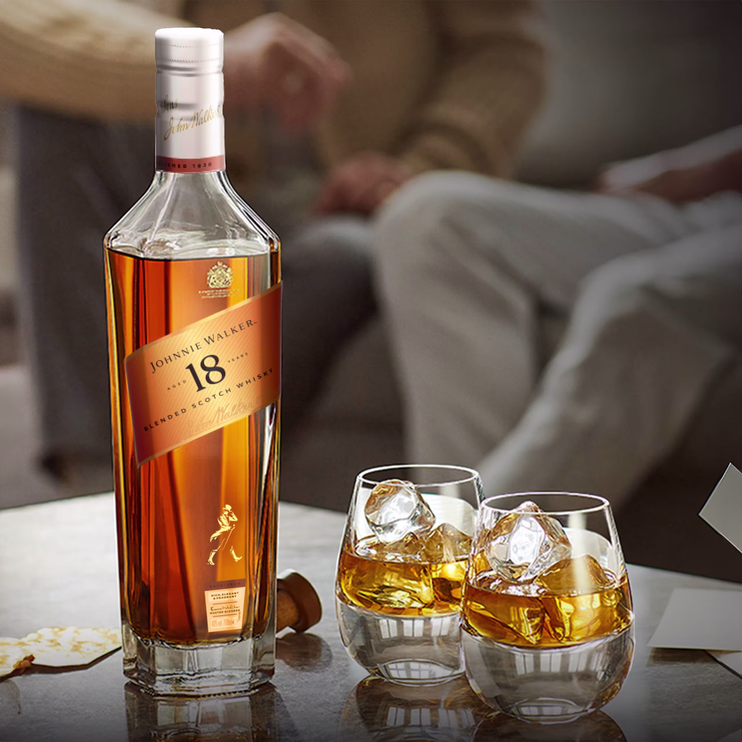 Láhev osmnáctileté míchané skotské whisky Johnnie Walker stojí na stole vedle dvou sklenic naplněných whisky a ledem; v rozmazaném pozadí sedí dva lidé.