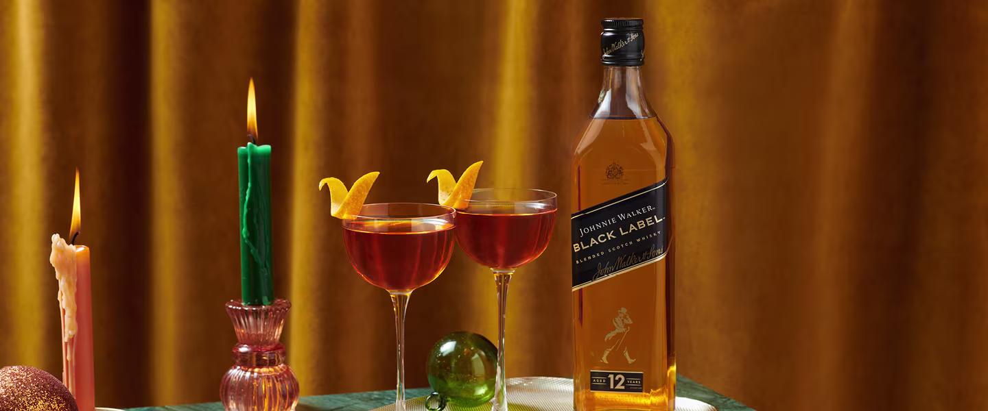 A bottle of Johnnie Walker Black Label whisky stands beside two cocktails with orange twists, a green candle, and a pink candle on a table with a yellow velvet curtain backdrop.