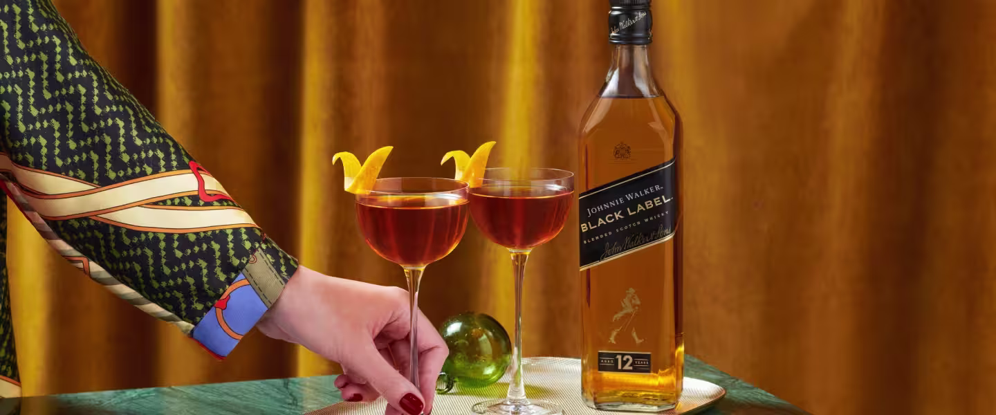 The Midnight Maple. A person wearing a patterned sleeve reaches for one of two cocktails on a table beside a bottle of Johnnie Walker Black Label. The glasses are garnished with orange peels, and a green glass sphere is nearby. 