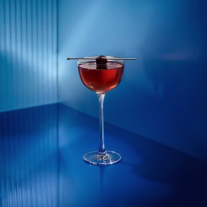 A cocktail with a cherry garnish on a skewer in a coupe glass, set against a sleek blue background.