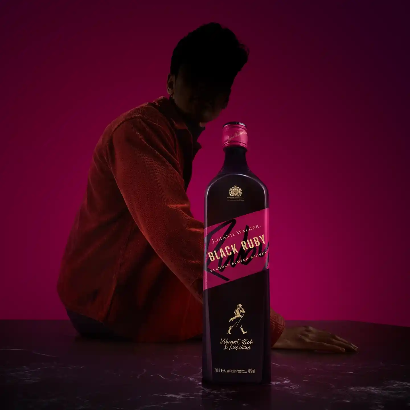 Una persona con una chaqueta roja posa de manera espectacular detrás de una botella negra con la etiqueta Black Ruby sobre una superficie oscura.